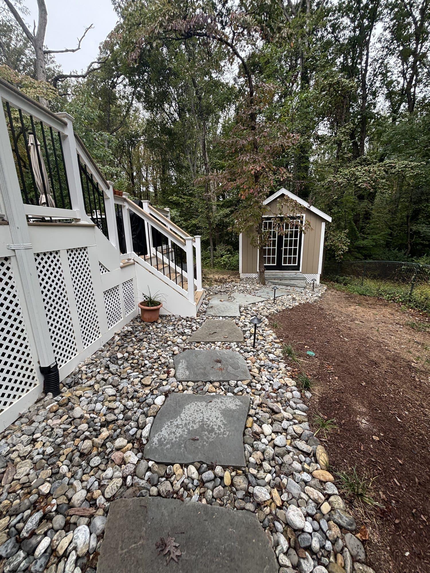 A finished backyard with a white deck, lattice skirting, and a stone-slab pathway to a board-and-batten outbuilding. Home addition project by Potts Brothers Construction, Arlington & Northern Virginia