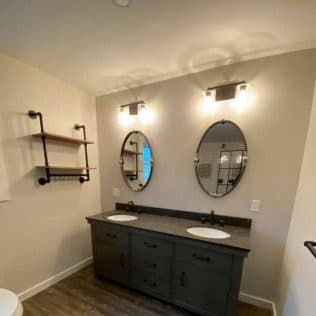 A finished bathroom with a dark double vanity, granite top, twin oval mirrors, pipe shelving, and wood-look flooring. Home addition project by Potts Brothers Construction, Arlington & Northern Virginia