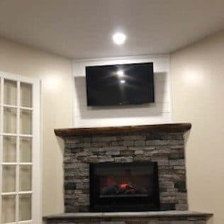 A finished corner stacked-stone fireplace with a reclaimed-wood mantel in a remodeled living room. Home addition project by Potts Brothers Construction, Arlington & Northern Virginia