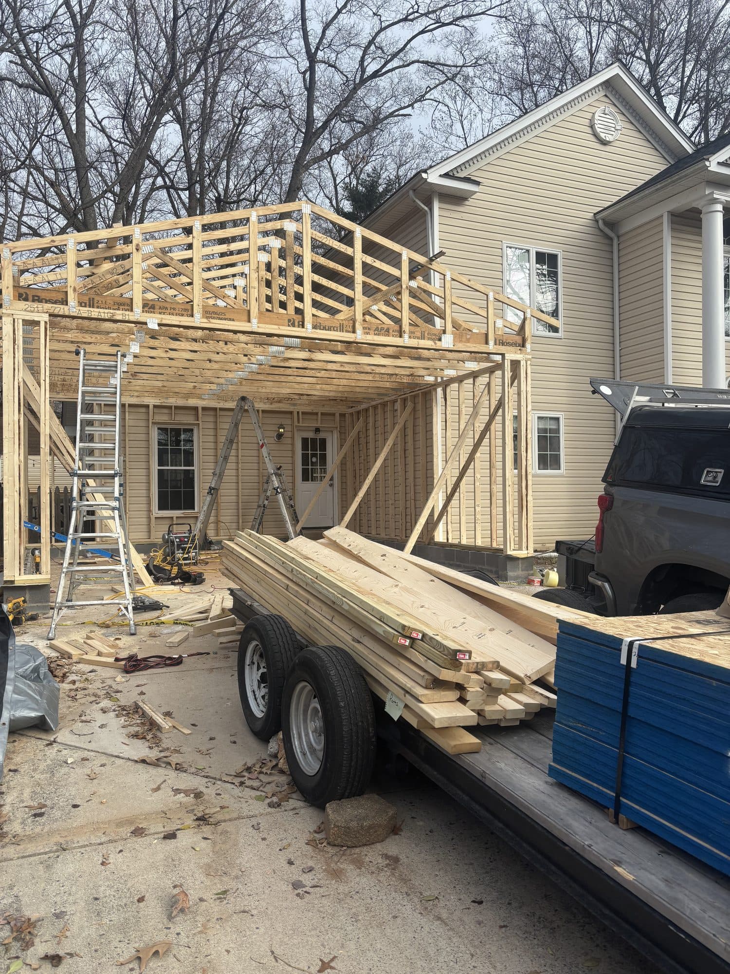 A framed two-story rear addition rises beside an existing sided home as a trailer delivers fresh lumber. Home addition project by Potts Brothers Construction, Arlington & Northern Virginia