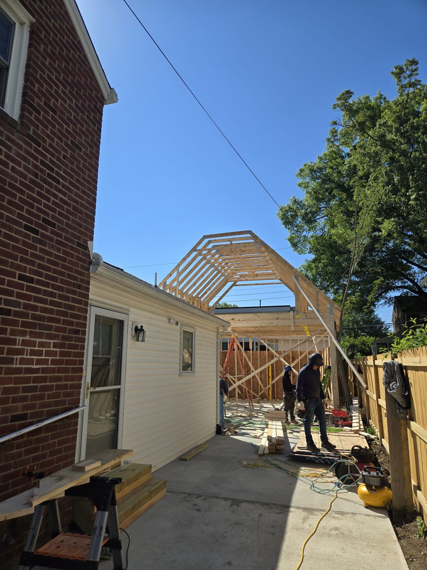 Workers frame the gambrel rafters of a rear addition attached to a brick home above a new concrete patio. Home addition project by Potts Brothers Construction, Arlington & Northern Virginia