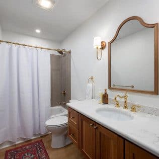 A remodeled bathroom with a wood vanity, marble top, brass fixtures, an arched mirror, and a tiled tub-shower. Home addition project by Potts Brothers Construction, Arlington & Northern Virginia