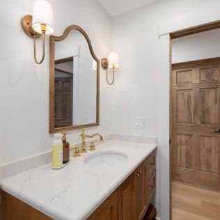 A remodeled bathroom with a wood vanity, marble countertop, arched mirror, and brass sconces and faucet. Home addition project by Potts Brothers Construction, Arlington & Northern Virginia