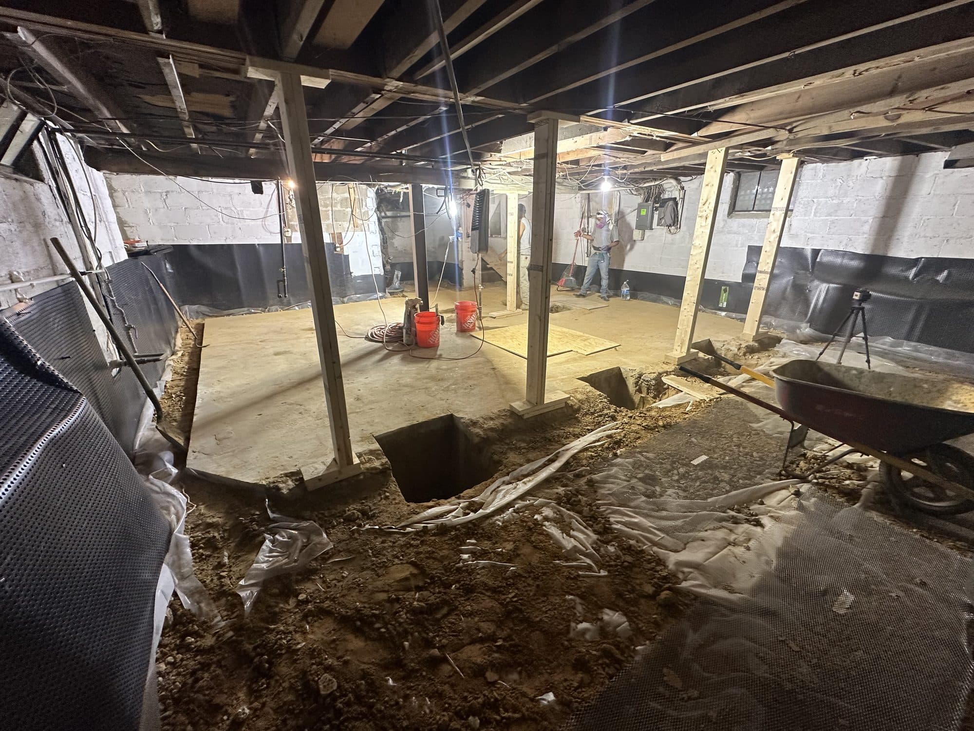 Basement interior mid-underpinning with open footing pits dug along the walls, temporary posts, wheelbarrow, and a drainage membrane on block walls. Foundation project by Potts Brothers Construction, Arlington & Northern Virginia